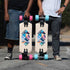 Two longboards are shown outdoors on a paved road, held upright by two partially visible people. The boards have a cream-colored deck with a colorful geometric graphic in the center, black trucks, and wheels in pink and turquoise blue.