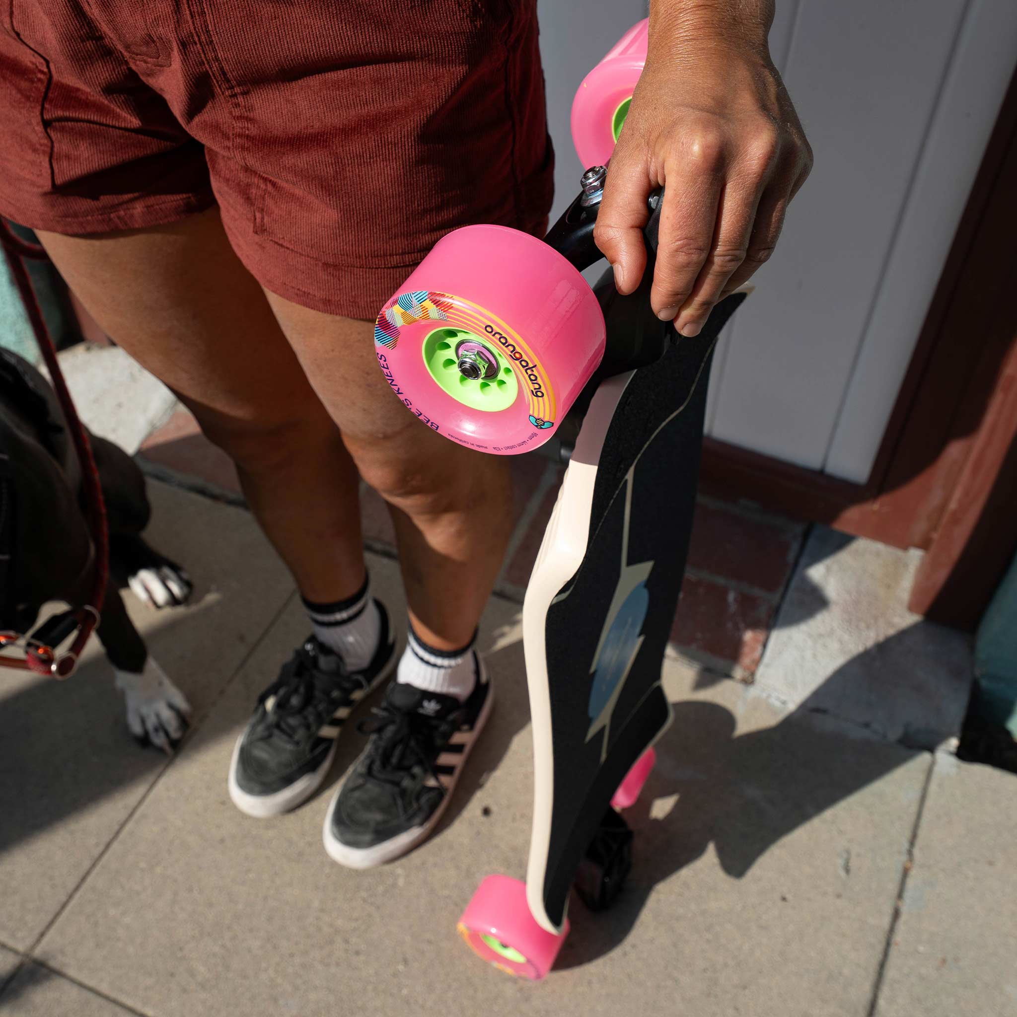 A person outdoors holding a longboard skateboard with bright pink wheels. The person is wearing rust-brown shorts, black and white sneakers, and white socks, standing on a paved sidewalk.