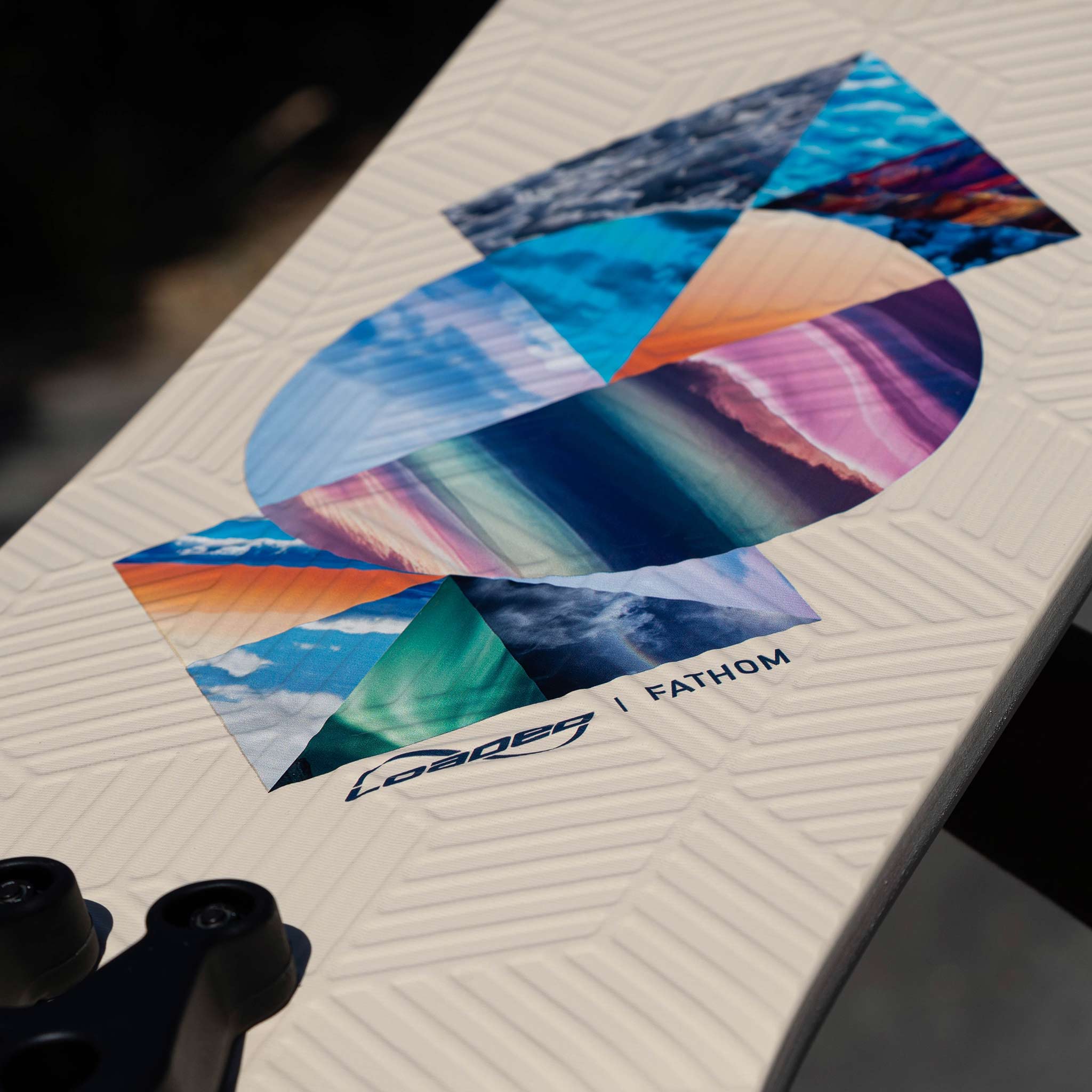 Close-up of a white textured deck with a colorful geometric graphic featuring the words 'FATHOM' and 'LOADED', with part of a black Zee Bracket visible in the lower left corner.