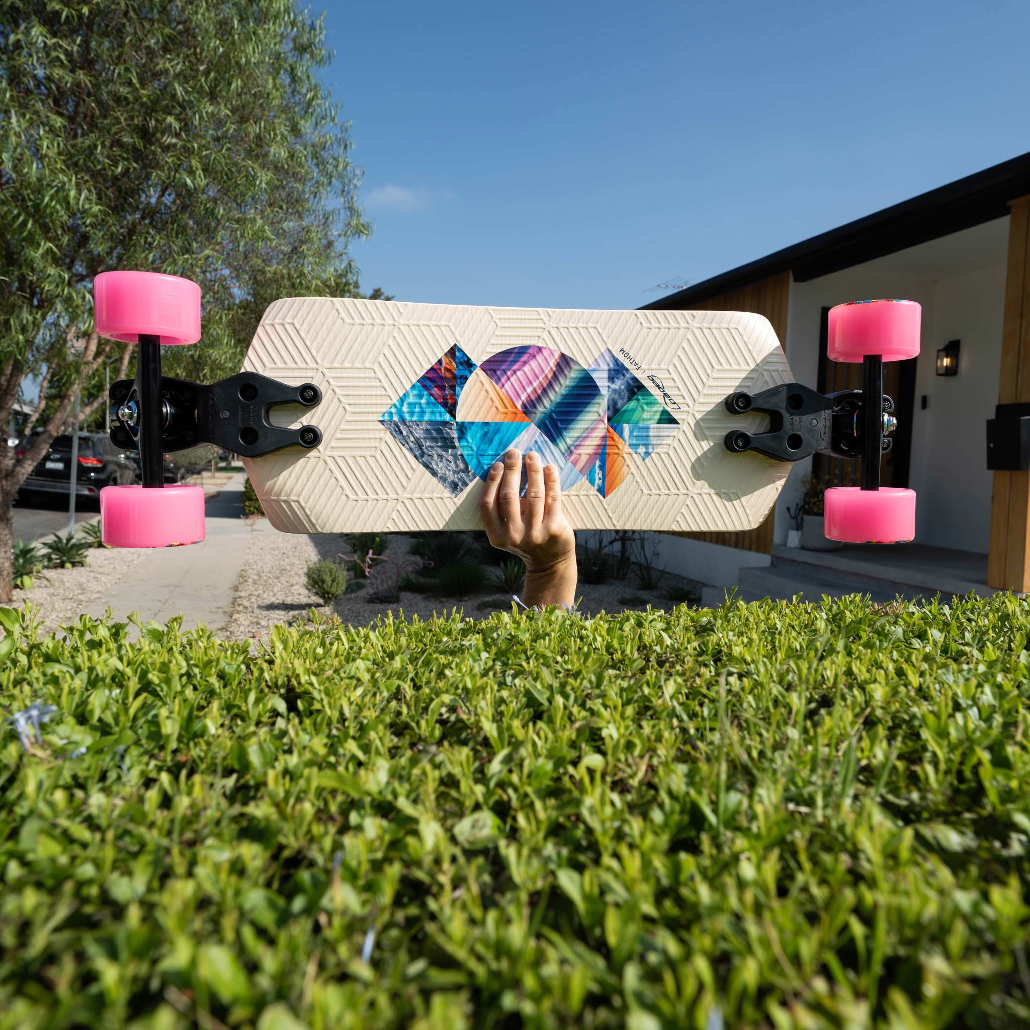 A person's hand holds a cream-colored longboard skateboard with a colorful geometric pattern on the deck and bright pink wheels, positioned in front of a green hedge and a modern house under a blue sky.