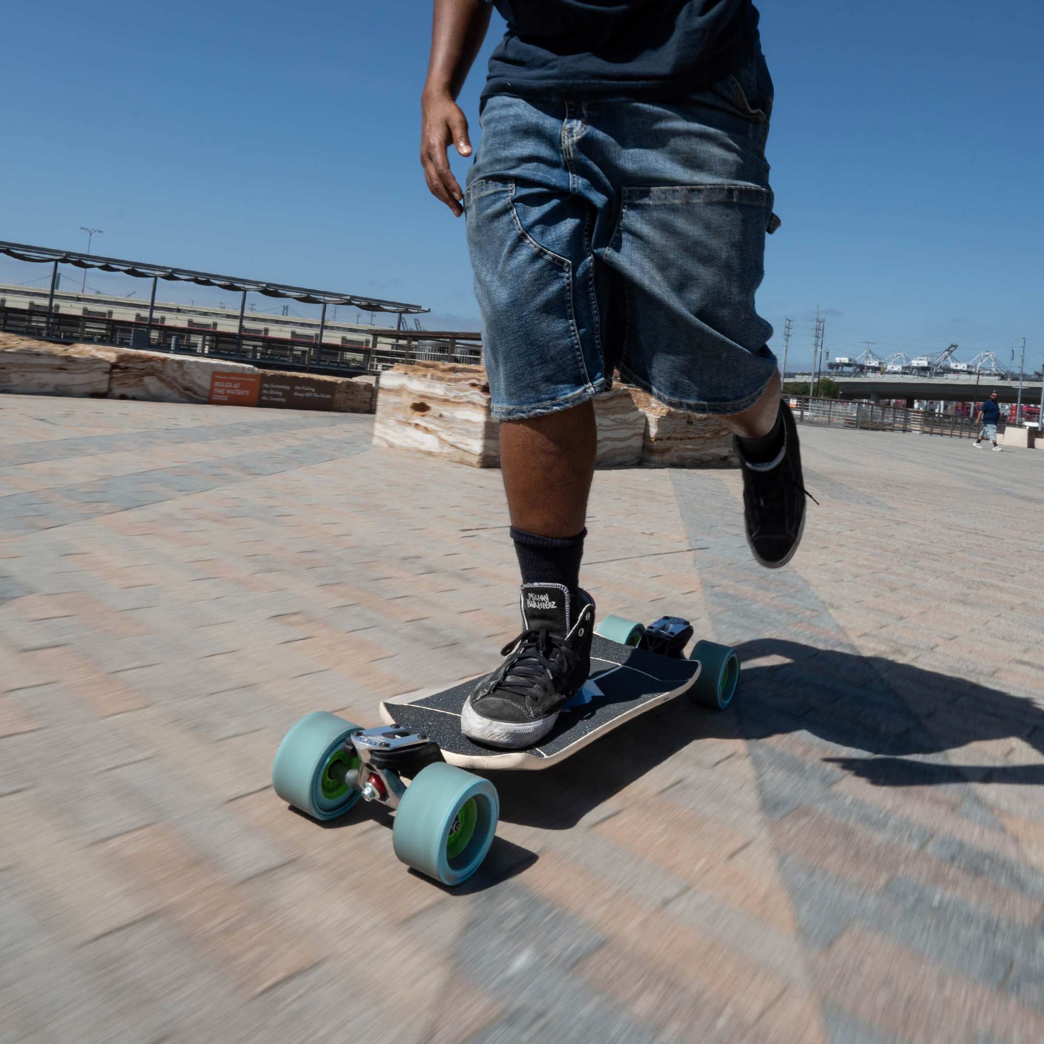 A person riding a longboard skateboard outdoors on a paved plaza under a clear blue sky. The skateboard has oversized blue wheels and a cream colored deck with black grip tape.