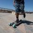 A person riding a longboard skateboard outdoors on a paved plaza under a clear blue sky. The skateboard has oversized blue wheels and a cream colored deck with black grip tape.