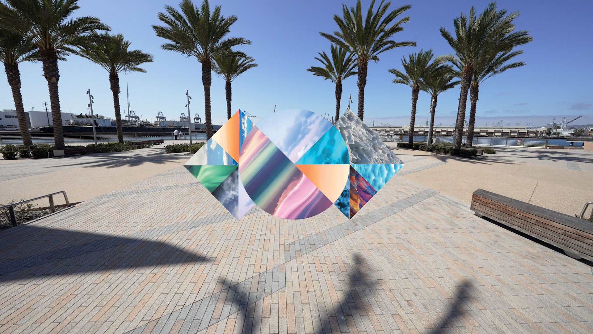A colorful abstract geometric wall art piece with overlapping circular and triangular panels in blue, pink, orange, green, and white, floating over on a paved waterfront plaza with palm trees and water in the background.