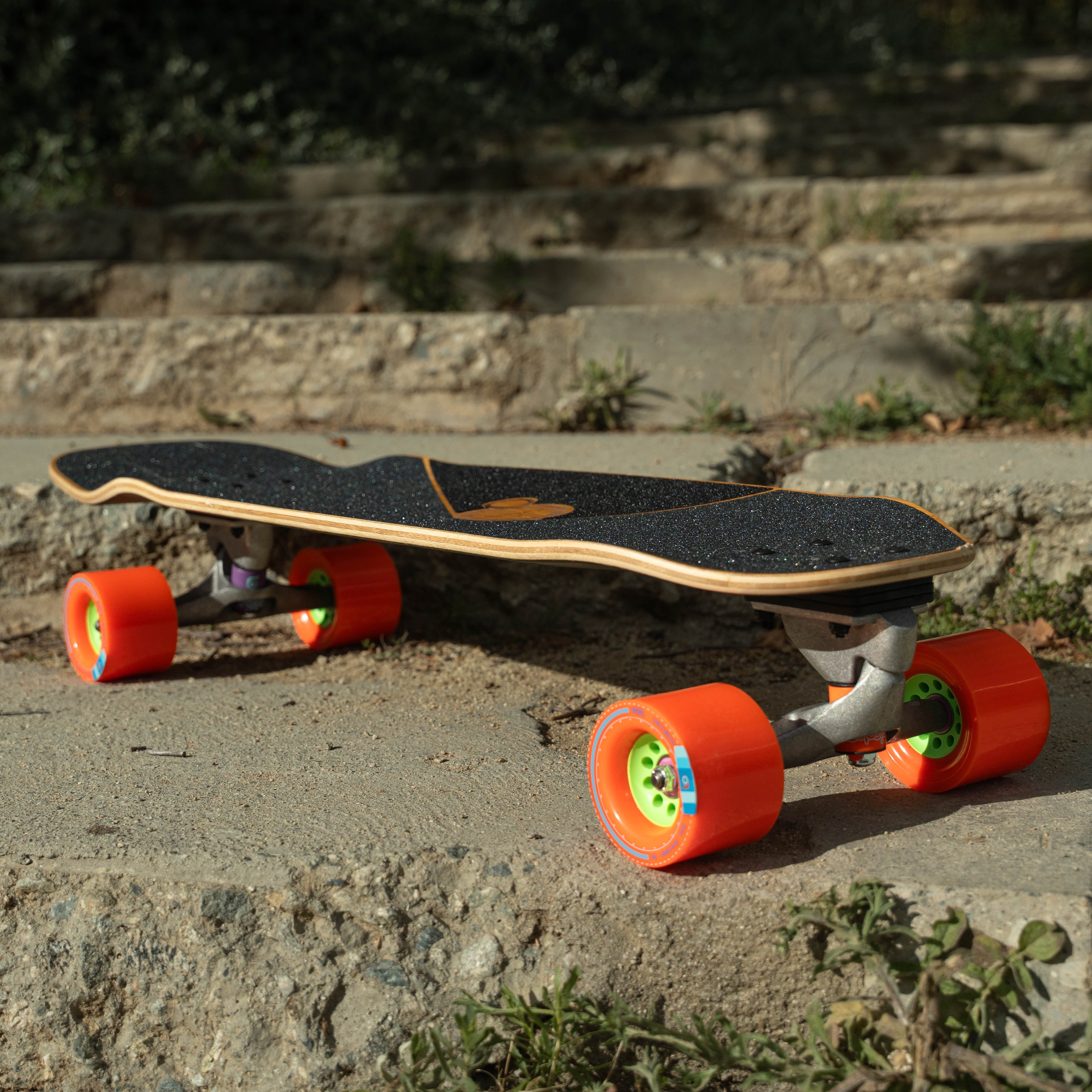 Loaded Omakase longboard with orange Orangatang Kegel wheels on a concrete surface with steps in the background