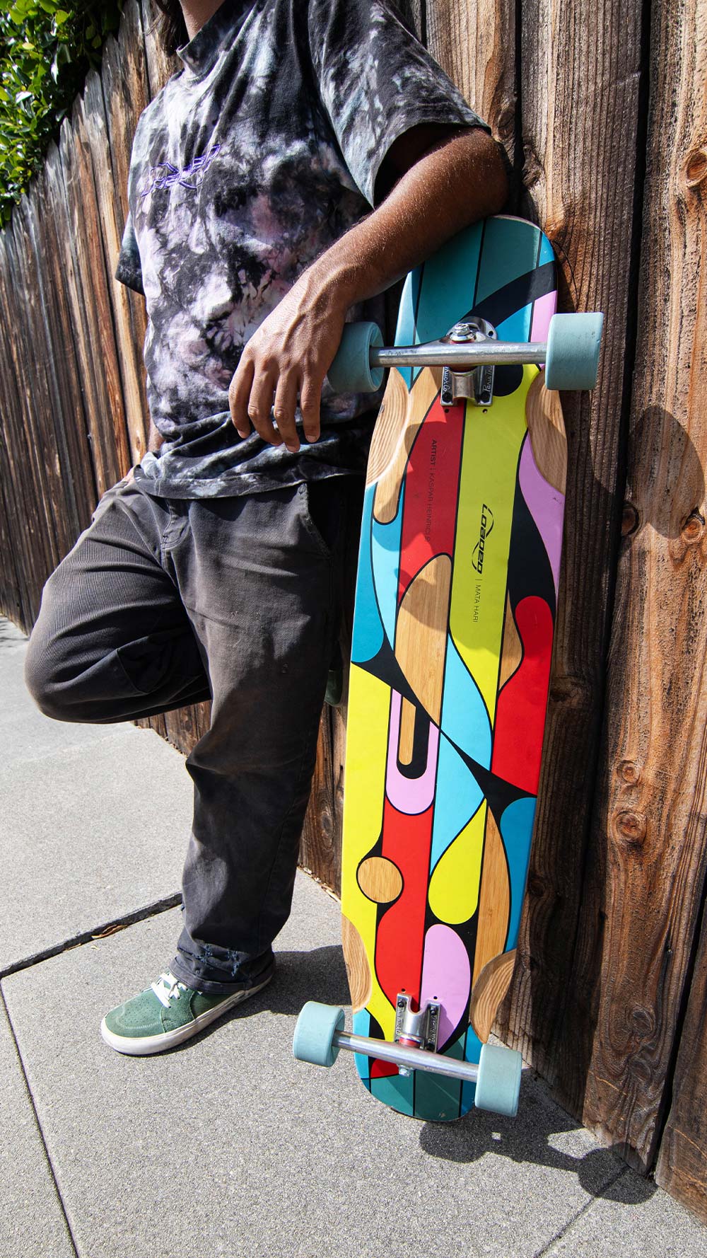 Man leaning against wooden fence with Loaded Mata Hari (Artist Series: Kaspar Heinrici graphic) dancing longboard skateboard