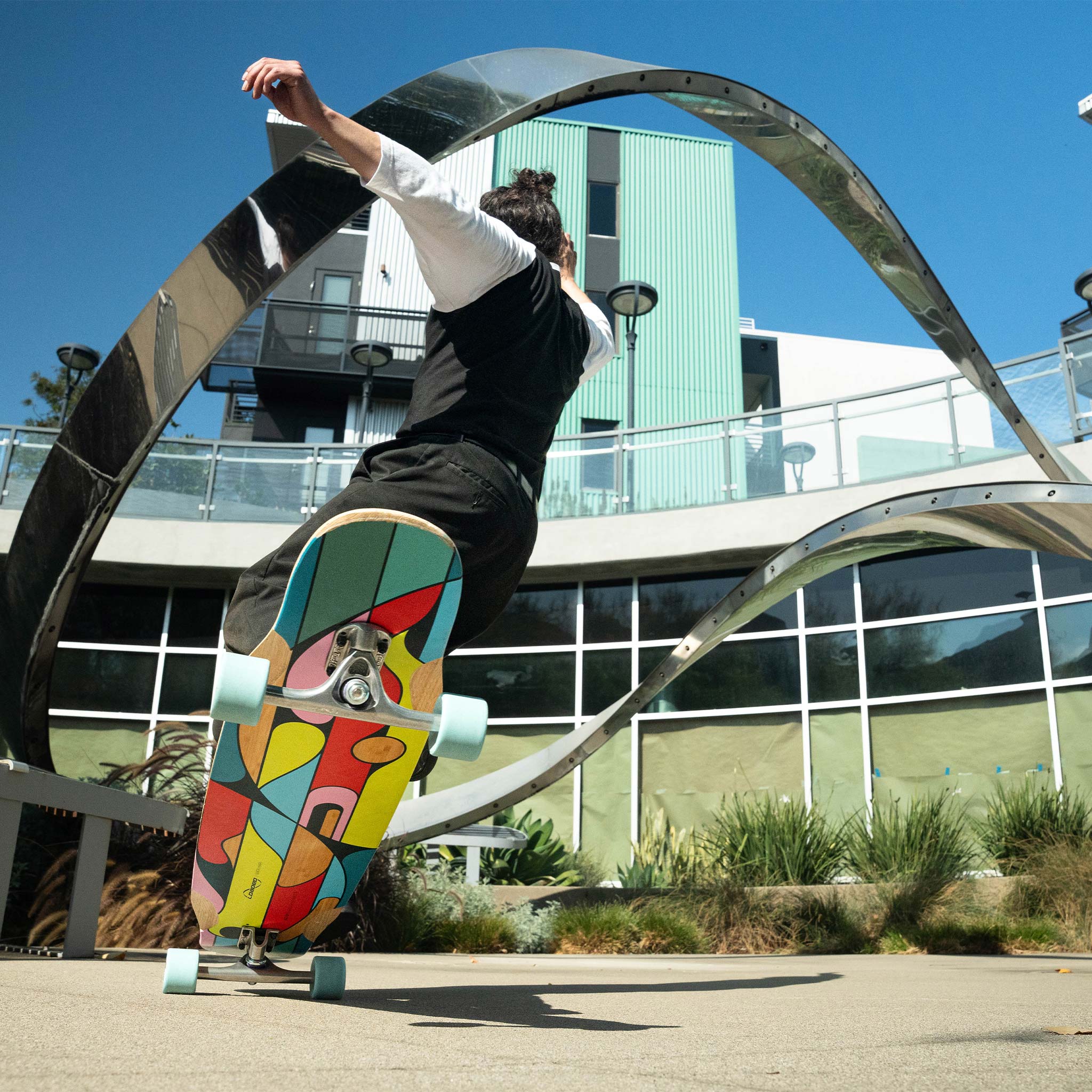 Skateboarder Mark Ponce performing a nose manual trick on a colorful Loaded Mata Hari (Artist Series: Kaspar Heinrici graphic) longboard skateboard in an urban setting with modern architecture.