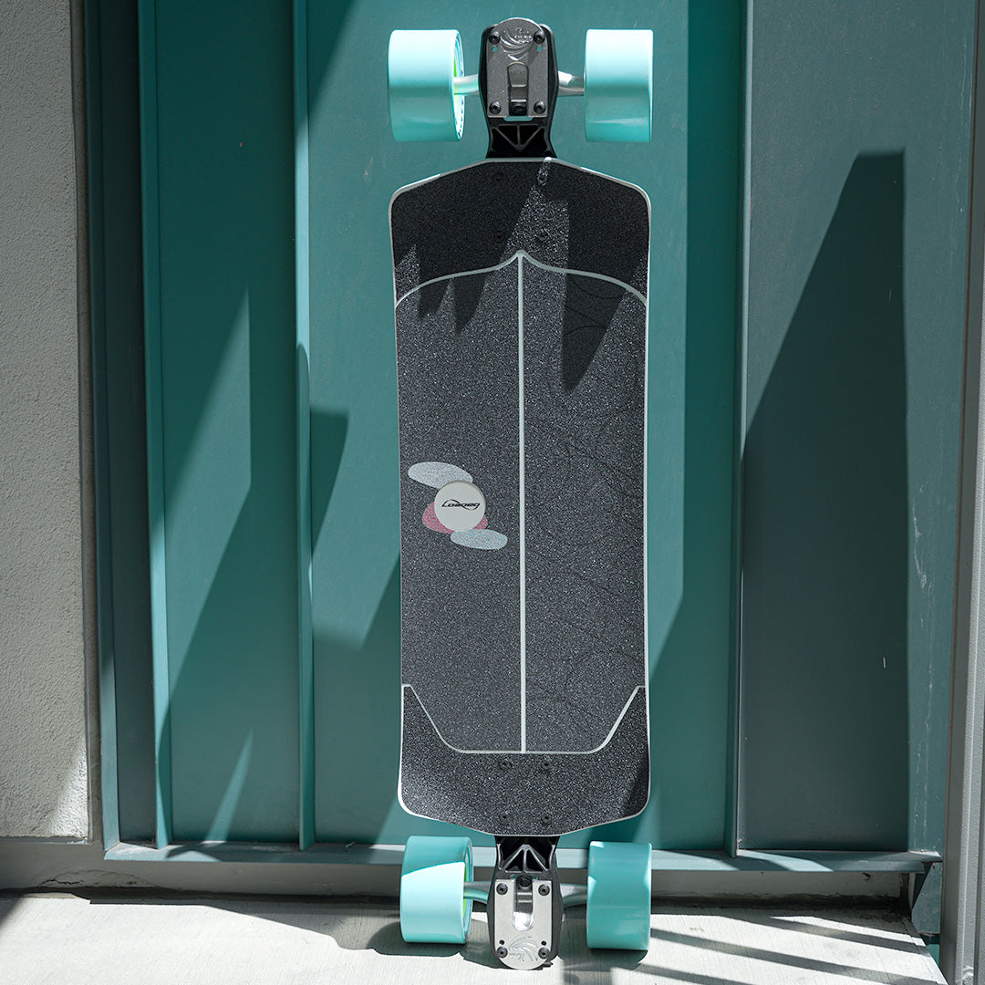 A longboard skateboard with a black textured deck, white graphic lines, and mint green wheels, photographed upright against a teal door with strong sunlight and shadows.