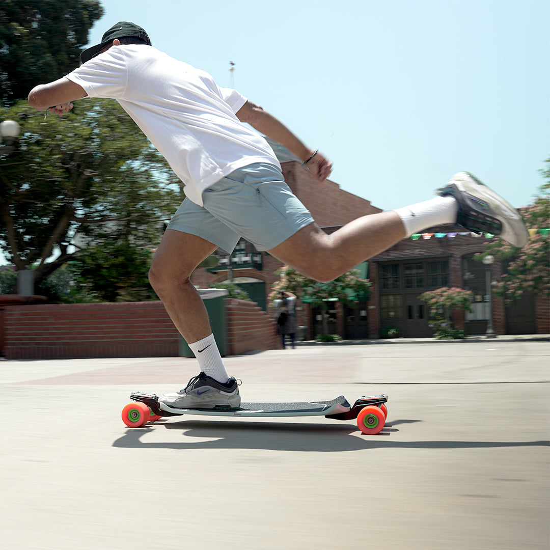 A man in a white t-shirt, light blue shorts, and a black cap is riding a double-drop longboard skateboard with bright orange wheels on a sunny outdoor paved area.